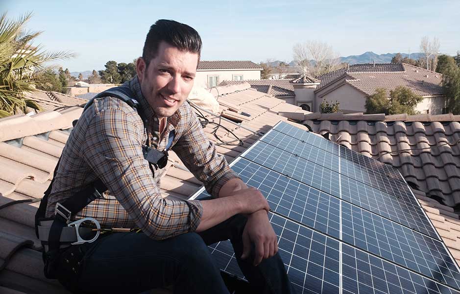 Jonathan Scott sitting on a tiled roof next to solar panels, wearing a safety harness
