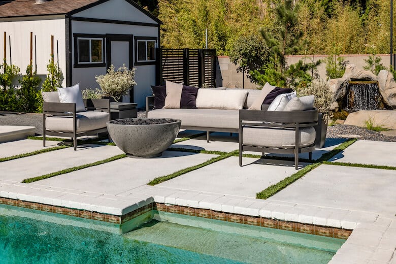 Poolside patio with modern outdoor seating, a round fire pit, and a small waterfall feature in the background