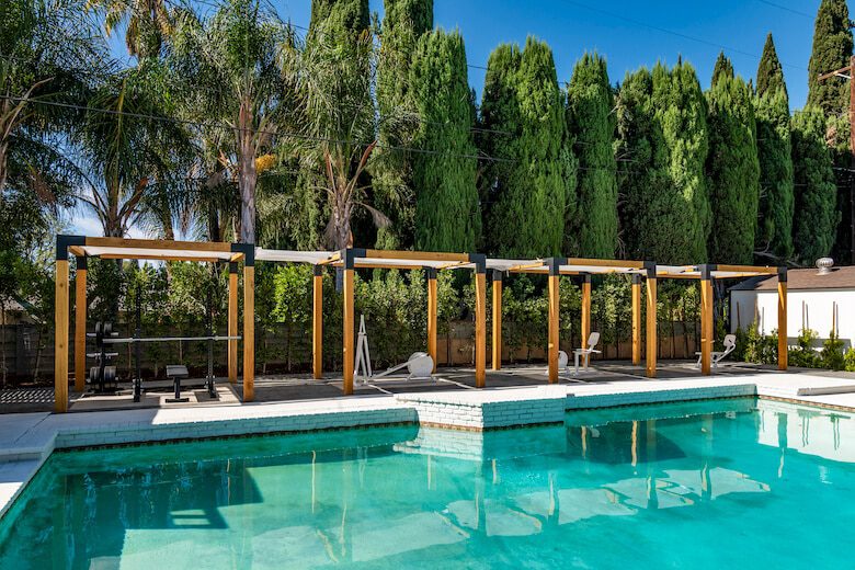 Outdoor pool area with wooden pergolas, tall greenery backdrop, and a mix of seating and gym equipment