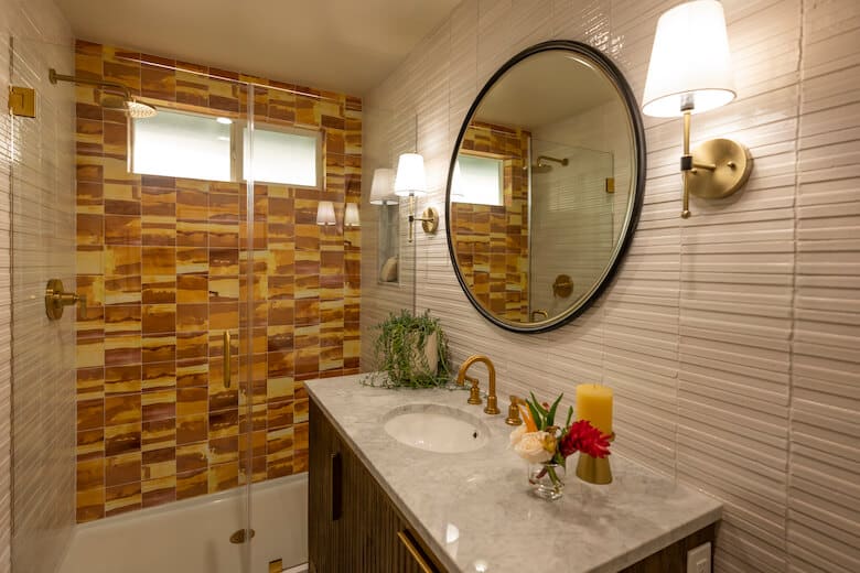 Bathroom with marble countertop, round mirror, gold fixtures, and yellow-brown tiled shower