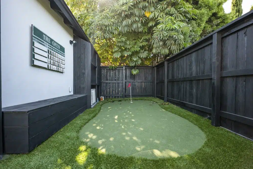Backyard putting green with black wooden fence, golf scoreboard, and surrounding trees