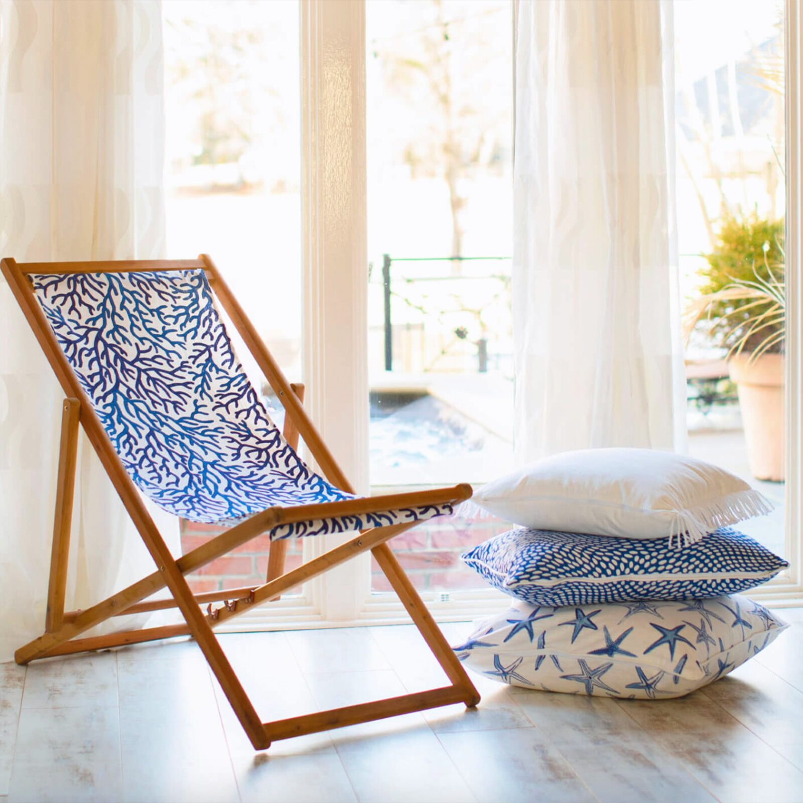 Beach-themed corner with wooden deck chair and stacked coastal print pillows by a window