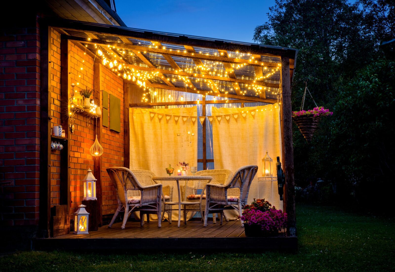 Outdoor patio with wicker furniture, string lights, lanterns, and flowers at dusk