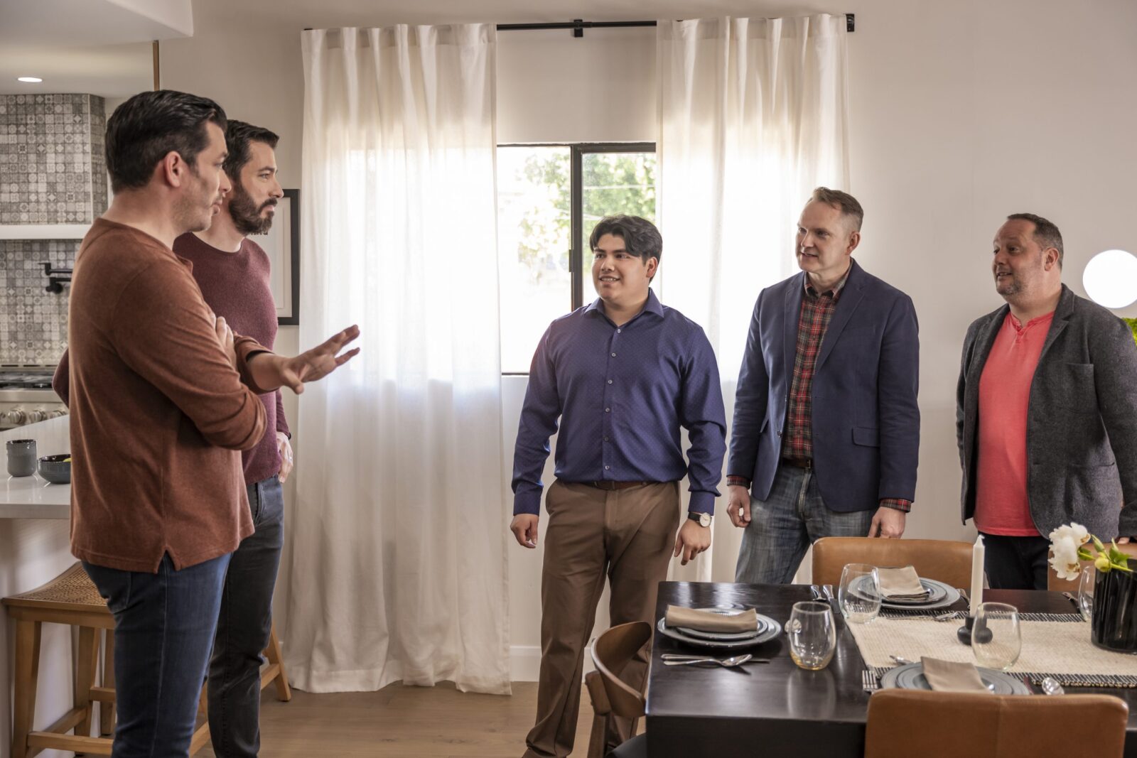 Group of five men talking in a modern dining area with set table, light curtains, and neutral decor