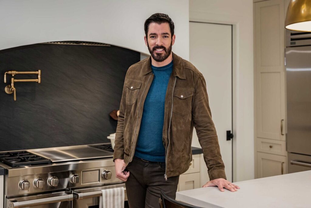 Drew Scott in a brown suede jacket standing in a modern kitchen with a black backsplash and brass pot filler, describing his tips for first-time homeowners