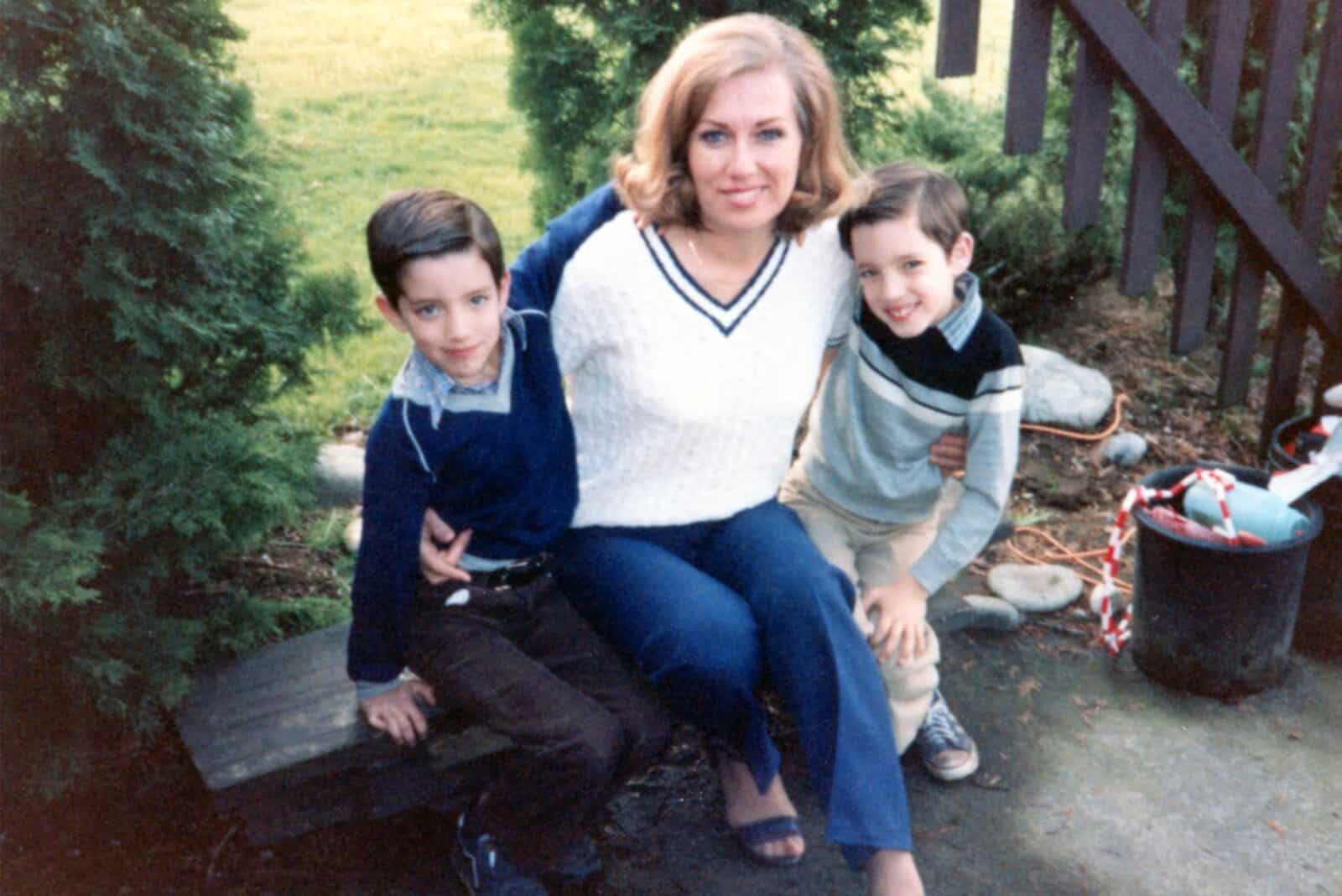 Drew and Jonathan with their mother sitting outdoors, posing for a family photo on a wooden bench