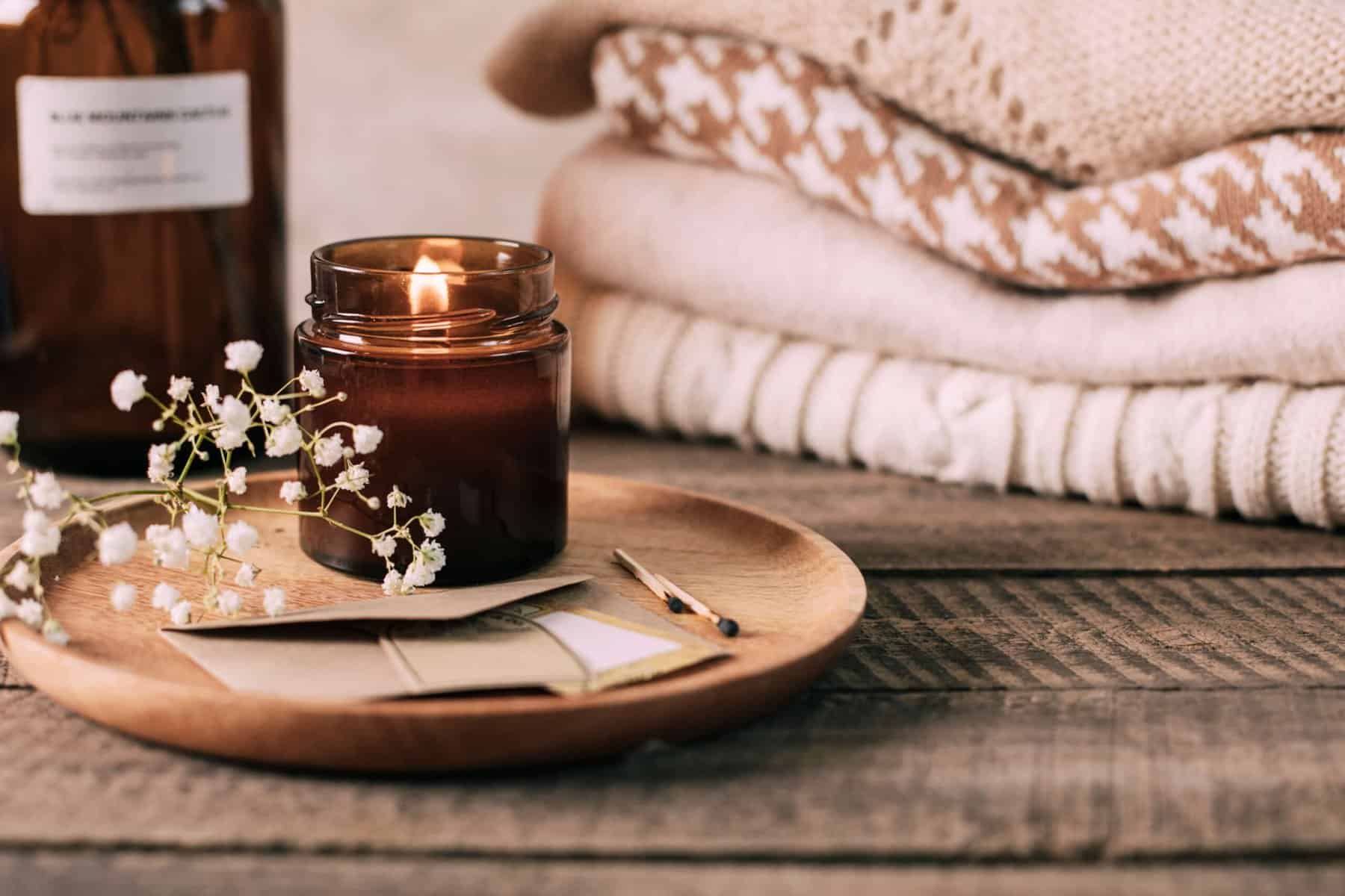 Cozy scene with lit amber candle, wooden tray, and folded neutral sweaters in the background