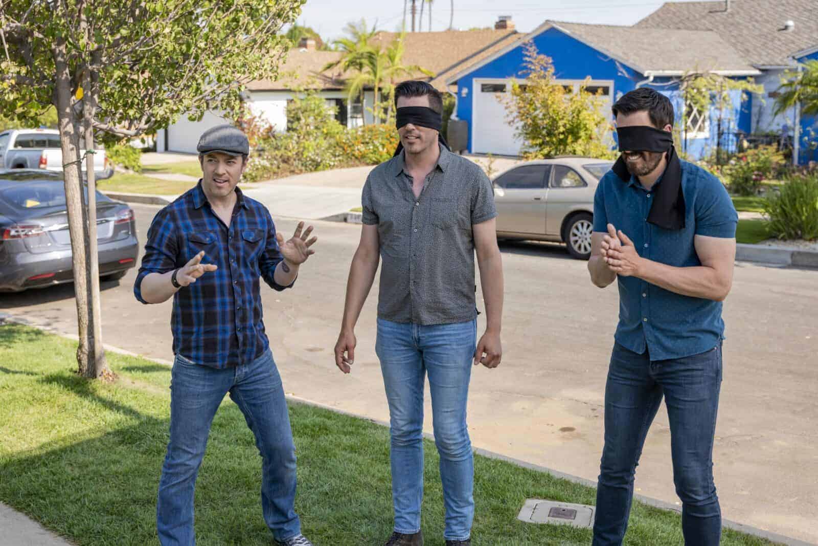 Man guiding Drew and Jonathan Scott, both blindfolded, outside a suburban home