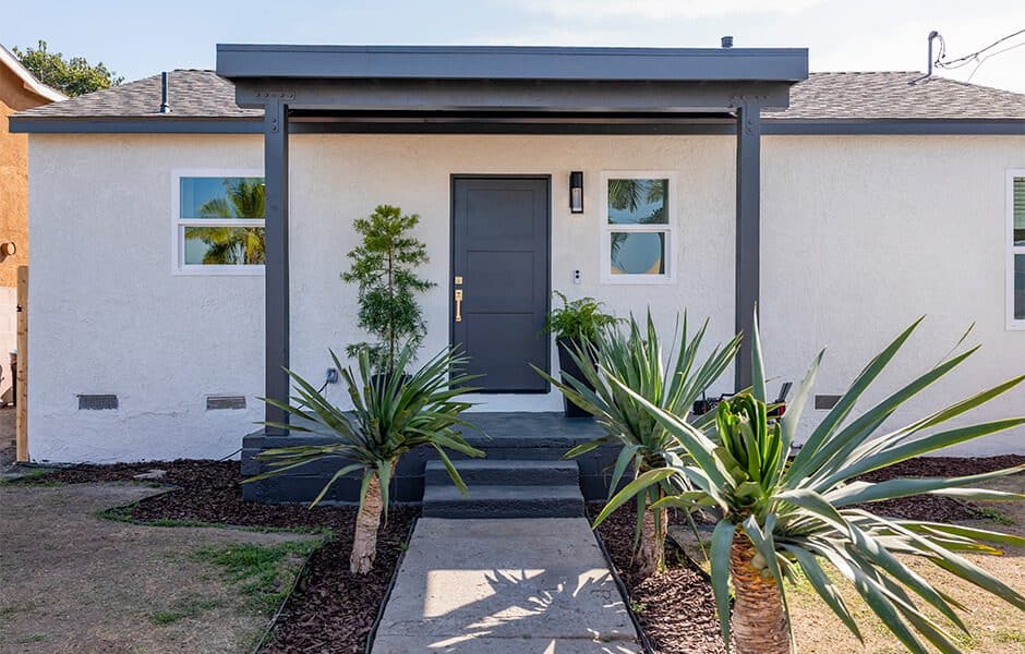 Small modern home with a dark front door, white exterior, and tropical landscaping