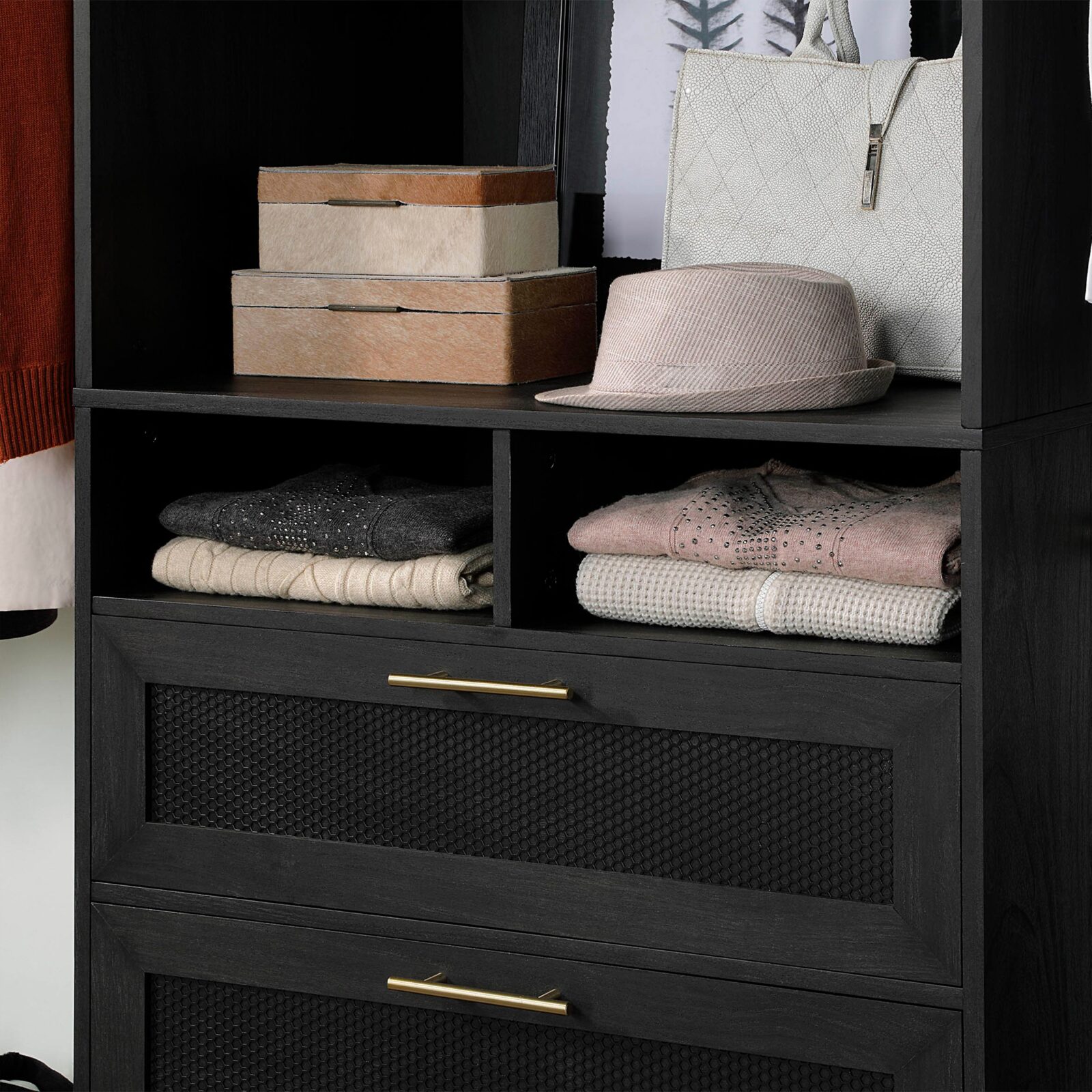 Black storage cabinet with gold handles, folded clothes, boxes, a hat, and a white handbag on display