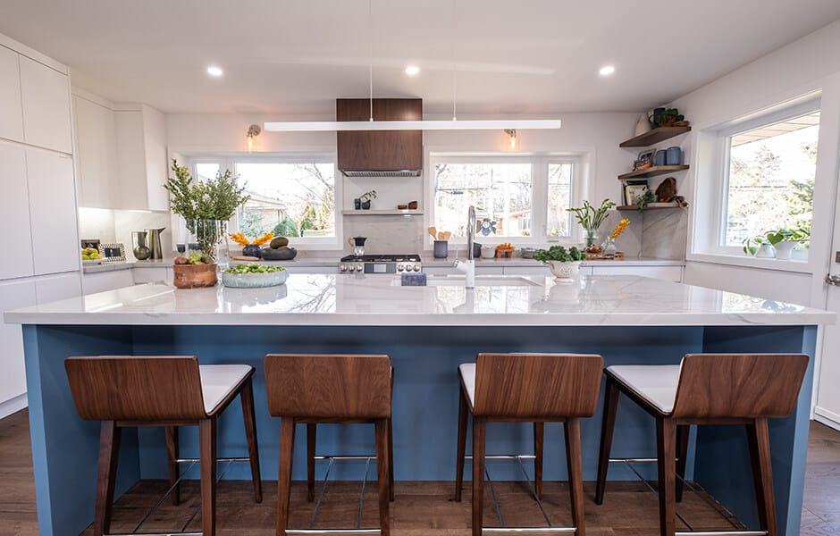 Bright kitchen with large island, blue base, wooden stools, and modern open shelving
