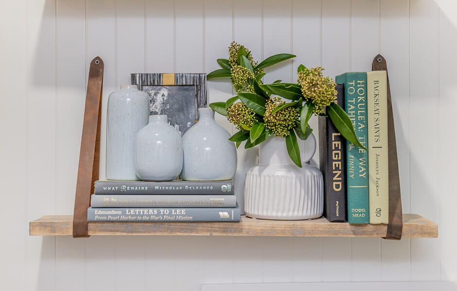 Wooden wall shelf with white vases, green foliage, and stacked books in a clean, minimal arrangement