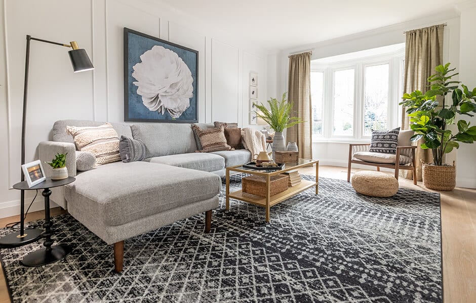 Bright living room with a gray sectional, large patterned rug, bay window, and indoor plants
