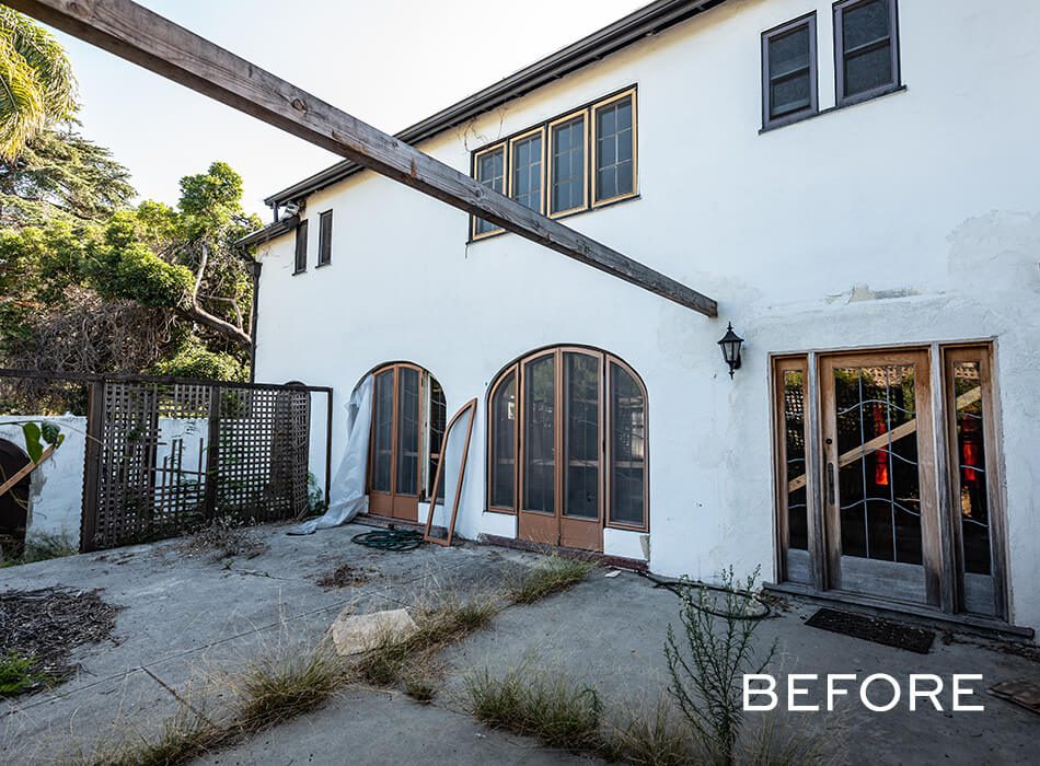 Weathered two-story house exterior with overgrown weeds, peeling paint, and wooden framed glass doors