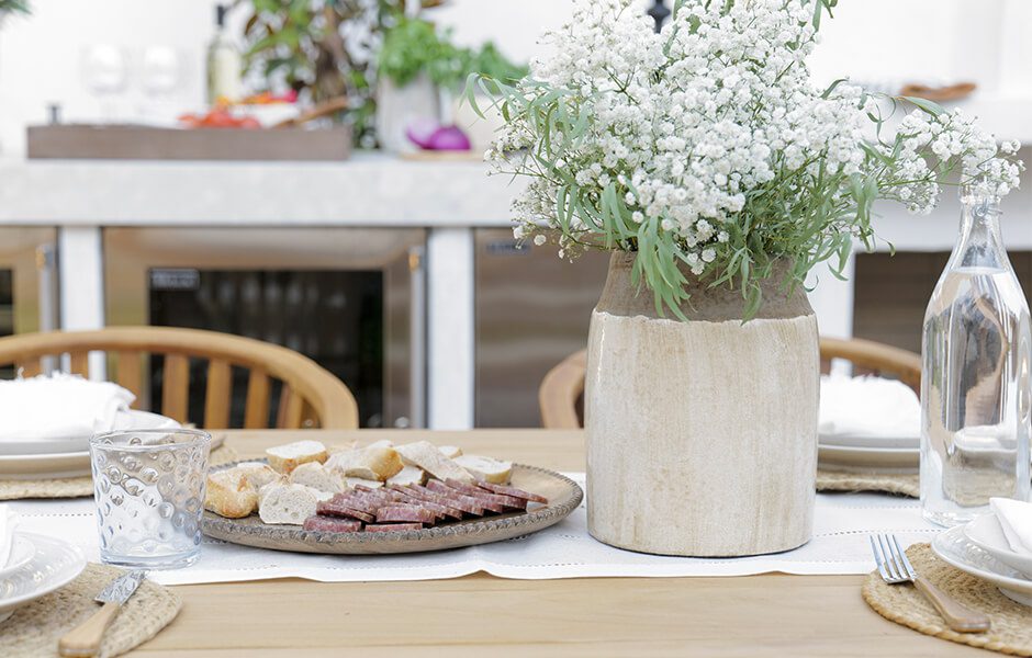 Close-up of outdoor dining table with a vase of white flowers and a plate of bread and sliced meat