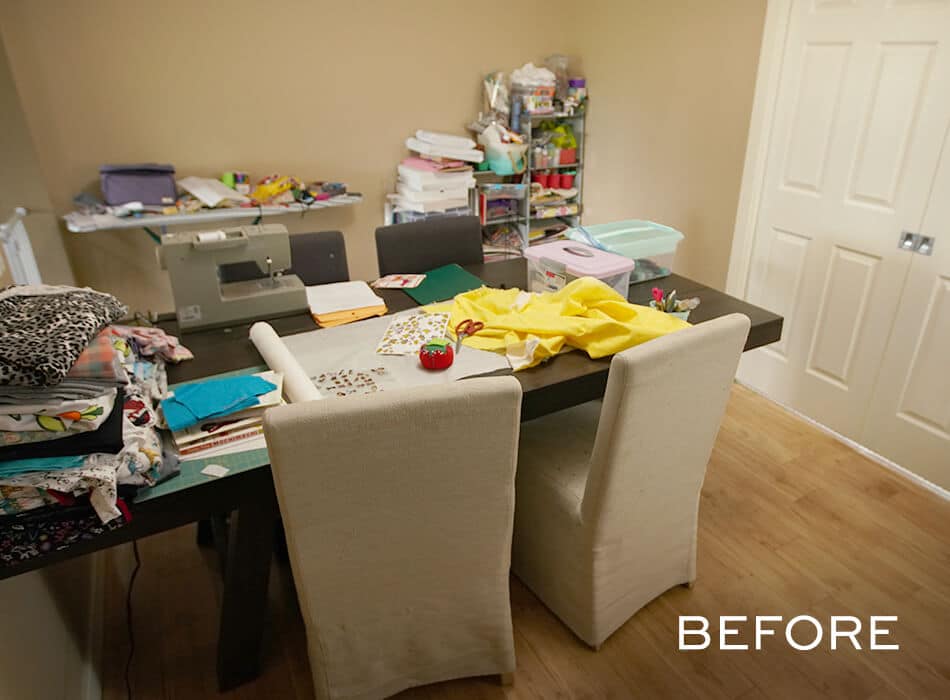 Craft room setup with sewing machine, fabric piles, storage bins, and supplies cluttering the table