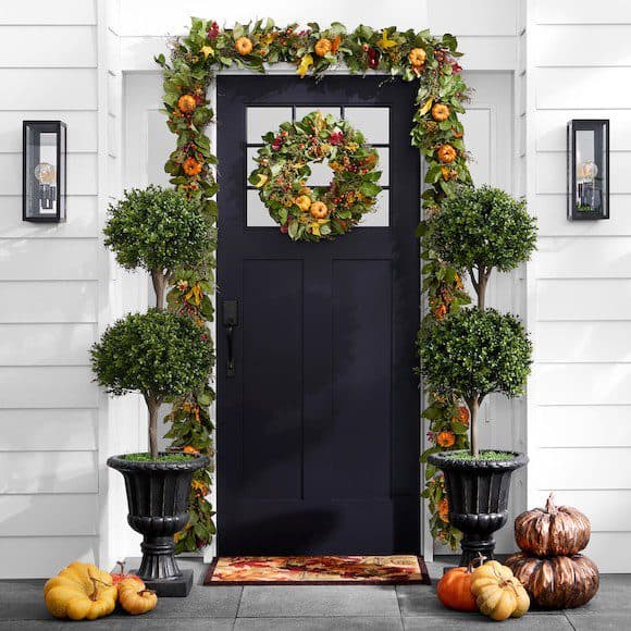 Front door decorated with a fall wreath, garland, potted topiary trees, and pumpkin