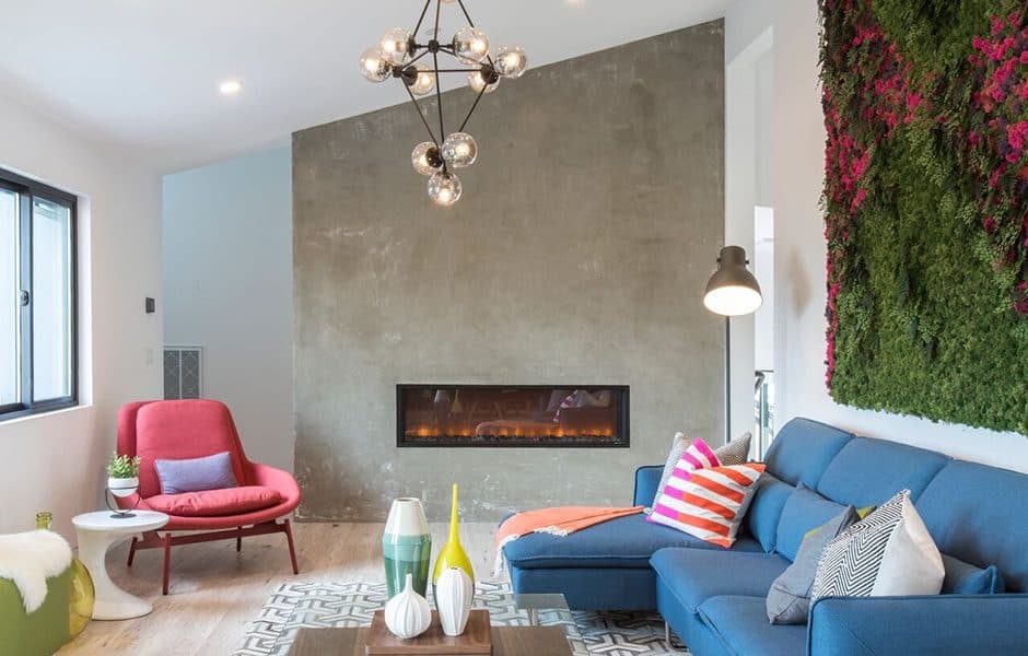 Modern living room with a blue sectional sofa, colorful throw pillows, a red accent chair, and a vertical green plant wall with pink flowers