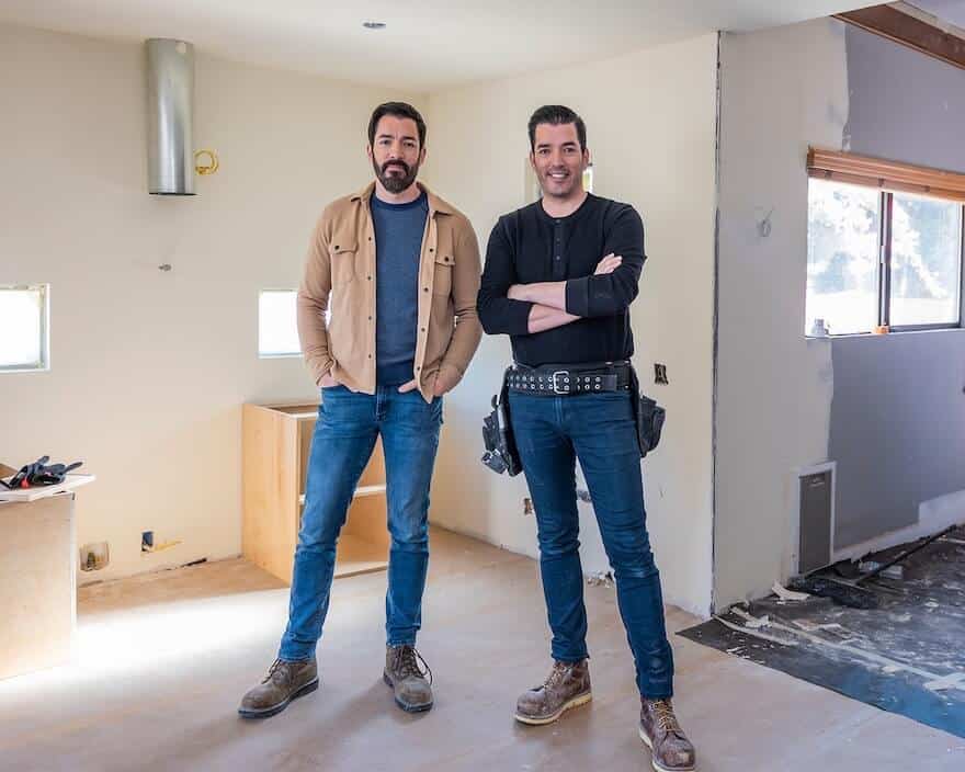 Drew and Jonathan standing in a partially renovated room, one in a tan jacket and the other in a black shirt with a tool belt