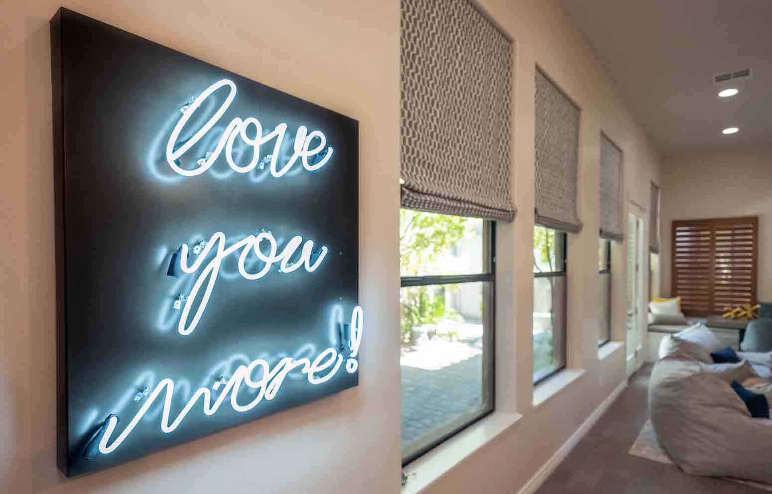 Neon sign that reads "Love you more!" on a wall next to windows with beige roller shades