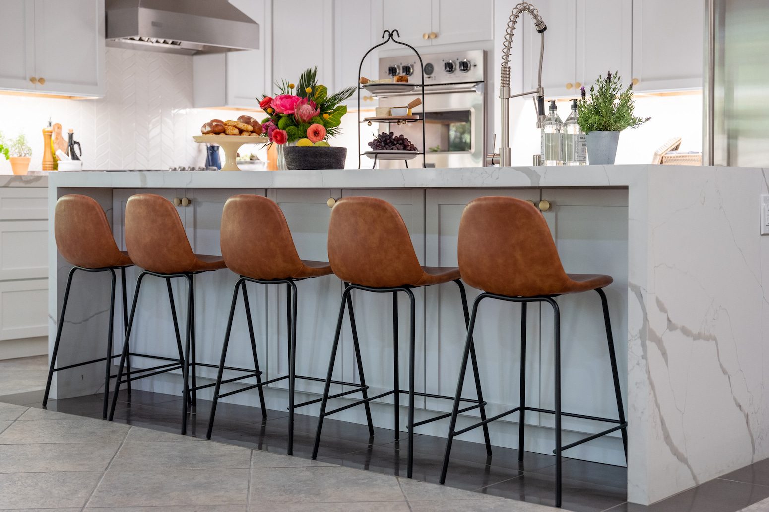 Modern kitchen with leather bar stools, a marble island, and a vase of fresh flowers and fruits on the counter
