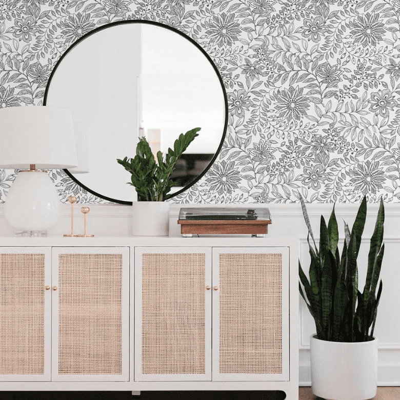 Entryway with floral wallpaper, round mirror, white console with rattan doors, potted plants, and a white lamp