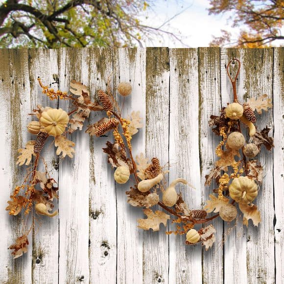 Fall garland with small pumpkins, pinecones, and leaves hanging on a rustic white wooden fence