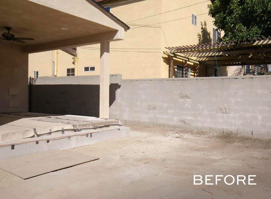 Before image of a backyard with a concrete wall, a covered patio area, and a gravel driveway