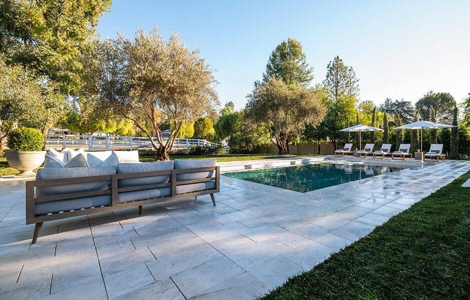Backyard pool area with lounge chairs, umbrellas, and cushioned outdoor sofa surrounded by trees