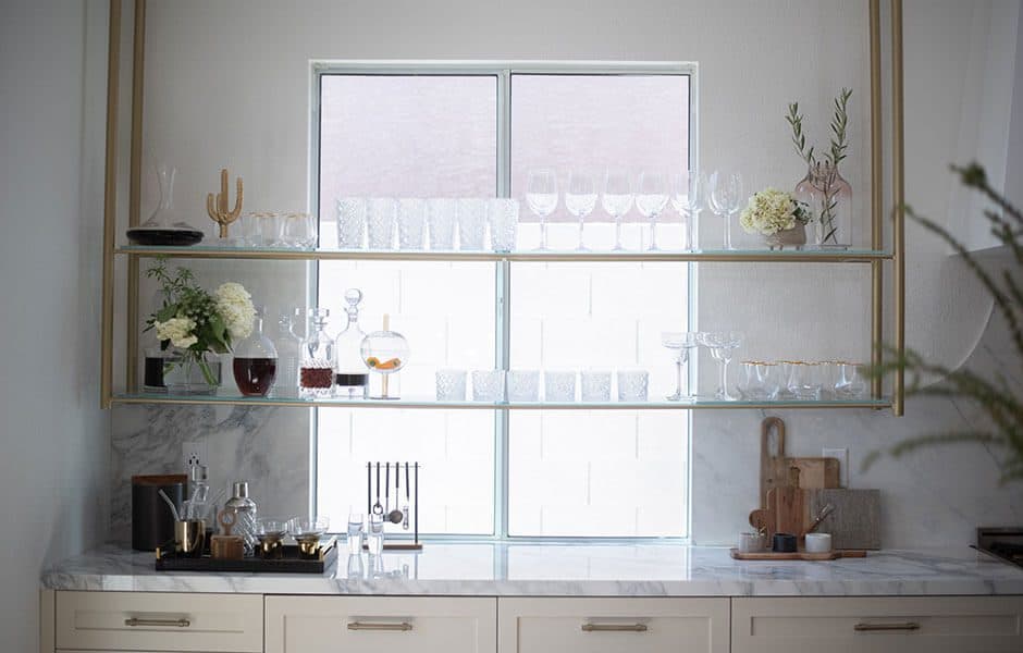 Home bar with marble countertops, gold-framed glass shelves, glassware, decanters, and decorative accents