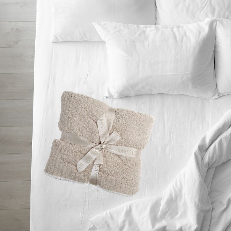 Beige plush blanket tied with a ribbon, placed on a neatly made white bed with soft pillows