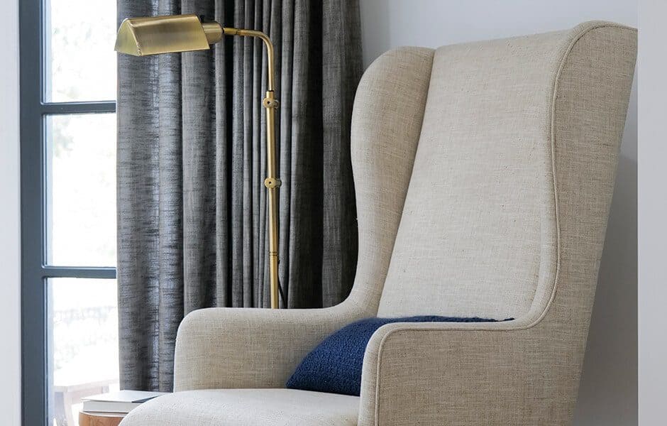 Beige wingback chair with a blue pillow, next to a brass floor lamp and a window with dark curtains