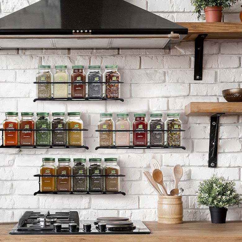 Kitchen spice rack display on a white brick wall with glass jars, wooden shelves, and a stovetop below