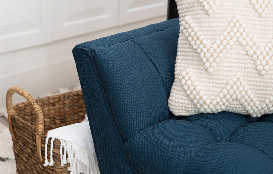 A close-up of a blue upholstered sofa with a white textured pillow, and a wicker backet in the background