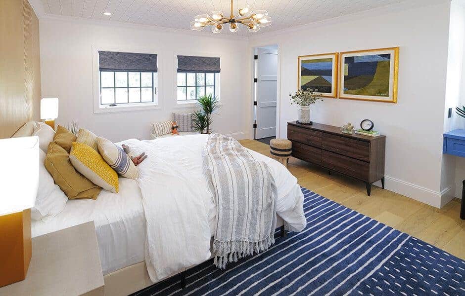 Modern bedroom with white walls, striped rug, wooden dresser, white bed with colorful pillows, and chandelier