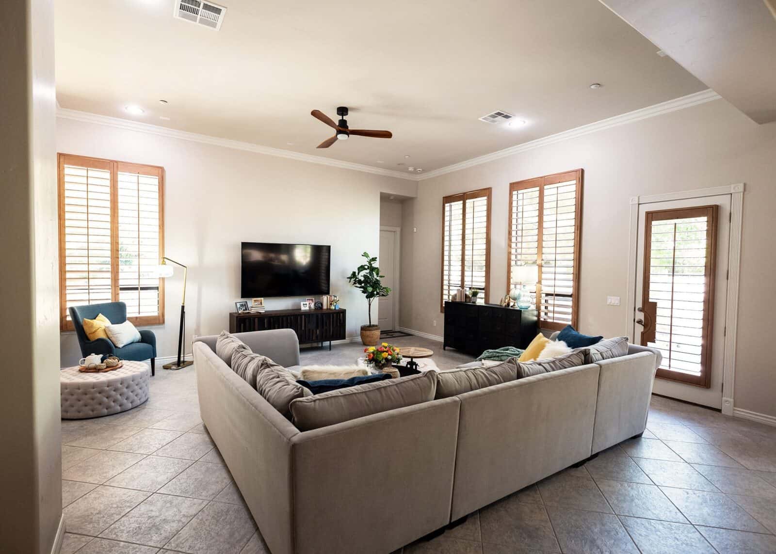 Living room with sectional sofa, coffee table, TV stand, ceiling fan, windows with shutters, armchairs, floor lamp, vases, and pillows