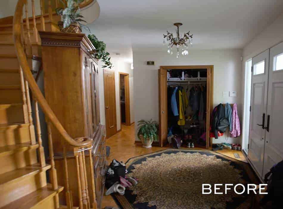 An entryway before renovation, featuring an open closet with jackets, a wooden staircase, a large area rug, and various items scattered on the floor