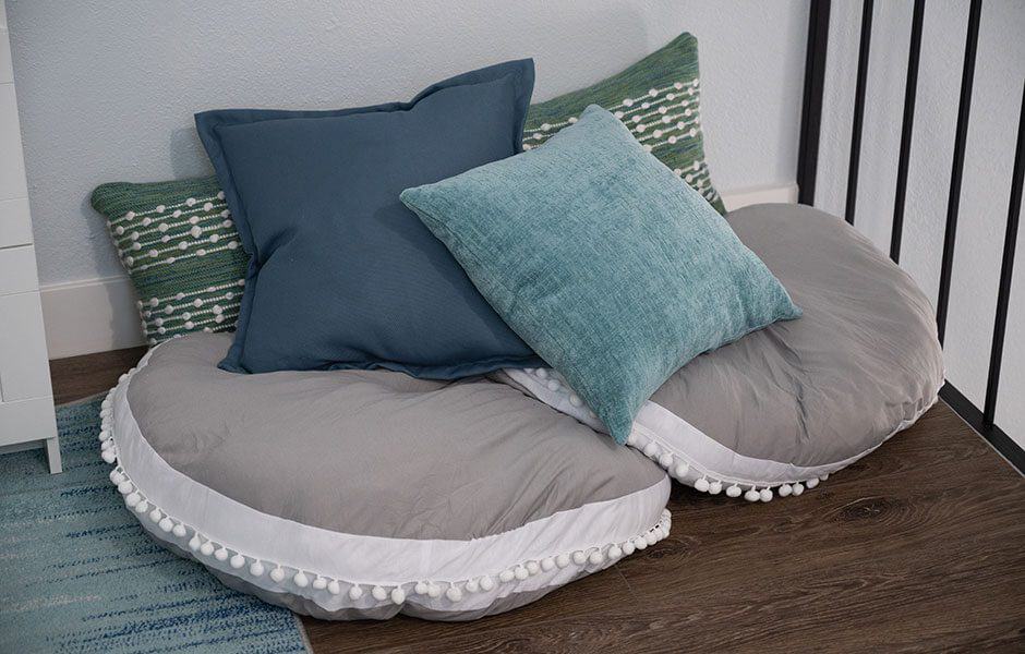 Two large, round floor pillows in a light gray color below blue, and green striped throw pillows