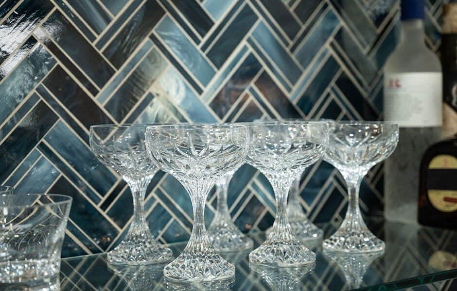 Crystal coupe glasses on a glass shelf with dark blue herringbone tile backsplash