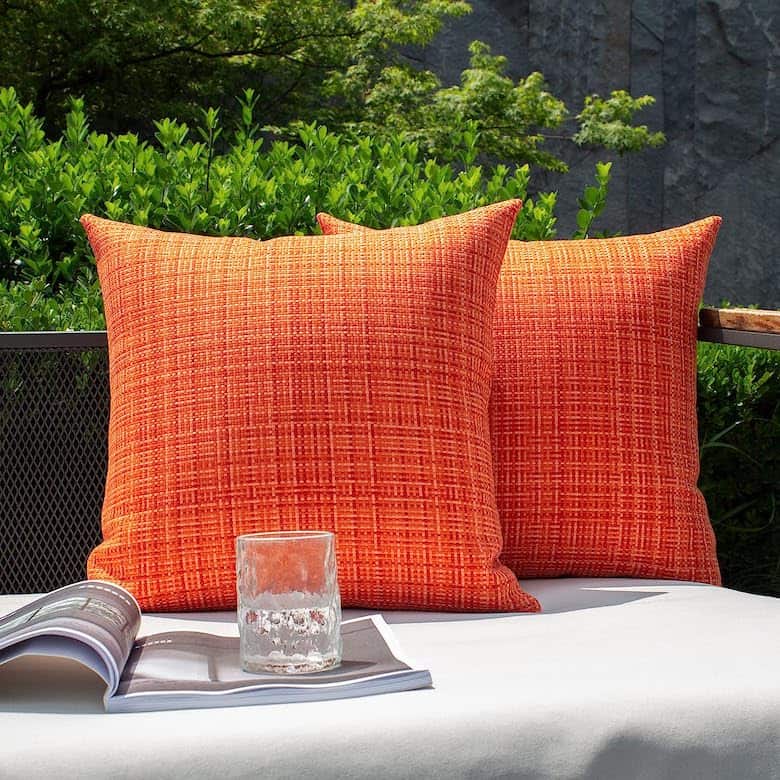 Two bright orange textured pillows on an outdoor bench with a glass of water and an open magazine