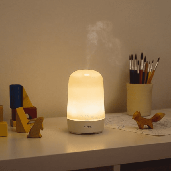 Lit Vitruvi diffuser on a desk with toys, brushes, and children's artwork