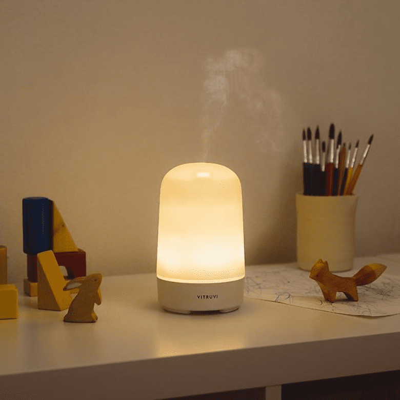 Lit Vitruvi diffuser on a desk with toys, brushes, and children's artwork