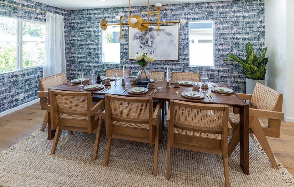 Modern dining room with wooden table, rattan chairs, patterned wallpaper, chandelier, flowers, and windows