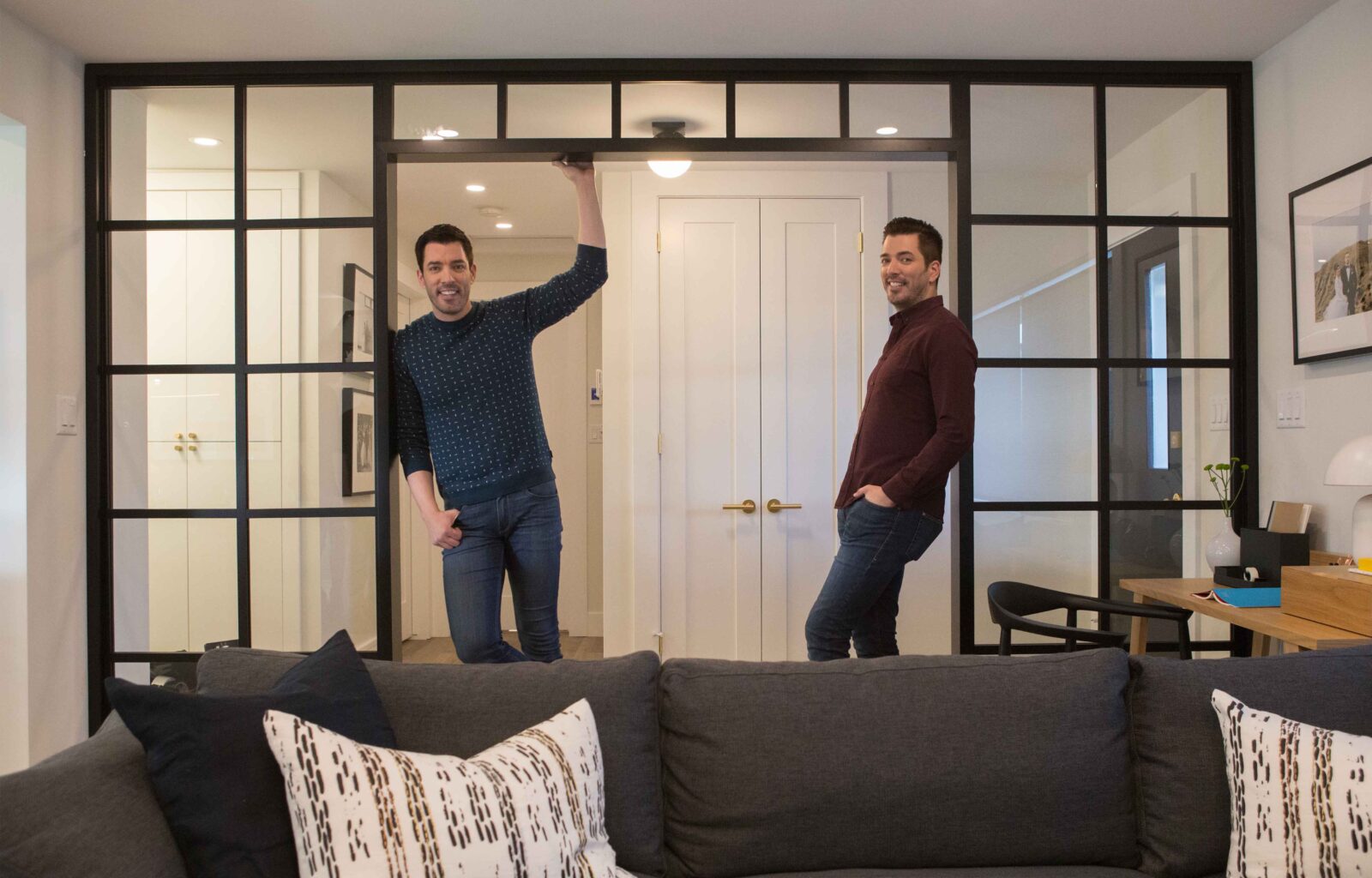 Drew and Jonathan Scott standing in a modern living room with black-framed glass doors