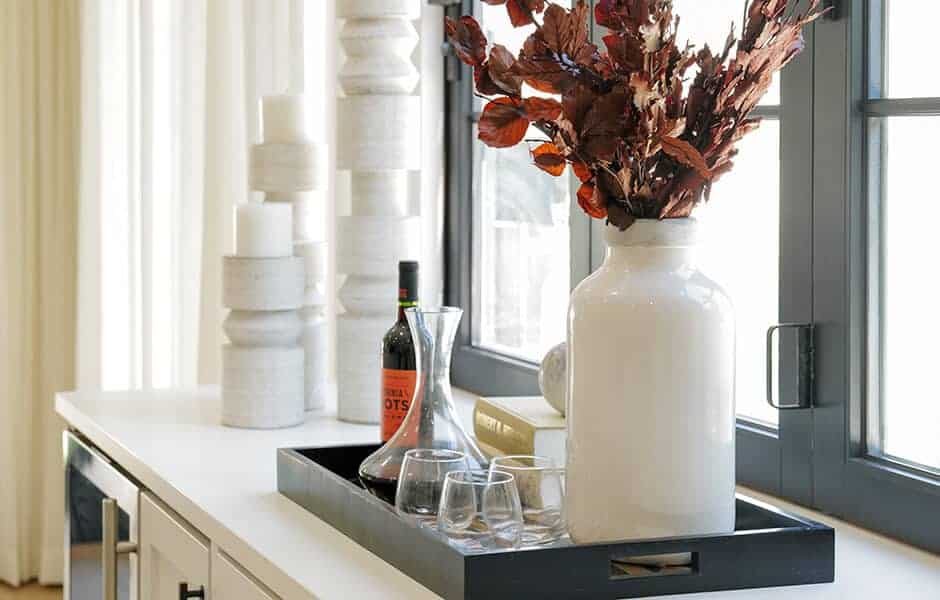 Decorative tray on a counter with a white vase of dried leaves, wine bottle, carafe, and glasses