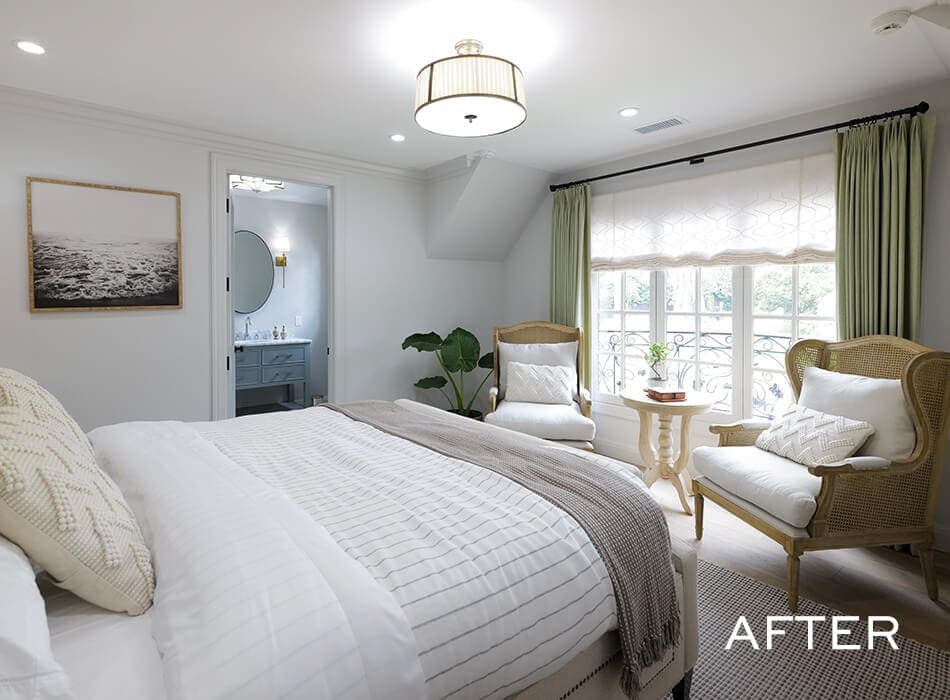 Bright bedroom with white bedding, wicker chairs by a window with green curtains, and a view into the bathroom
