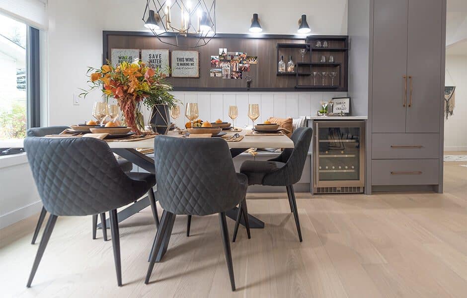 Elegant dining area with quilted gray chairs, light wood table, and a built-in bar with dark shelving and modern lighting