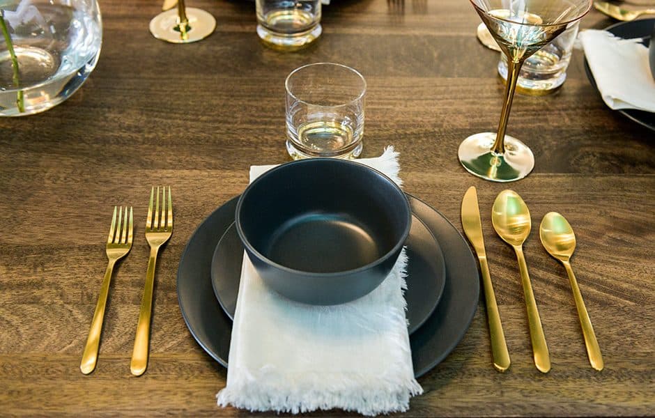 Elegant table setting with matte black dinnerware, gold flatware, and a white fringed napkin on a wooden table