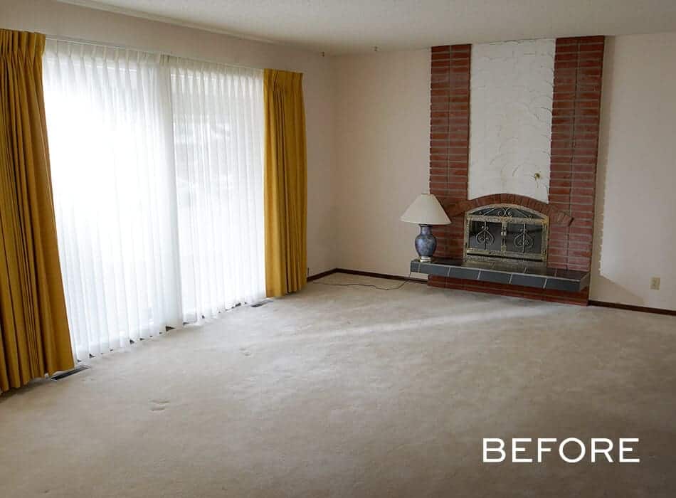 A before image of an empty dated living room with a brick fireplace, yellow curtains, and a carpeted floor
