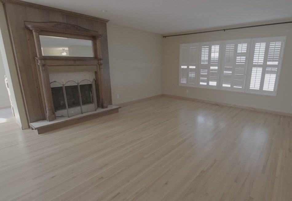 Empty living room with wooden fireplace, light hardwood flooring, and large windows with shutters